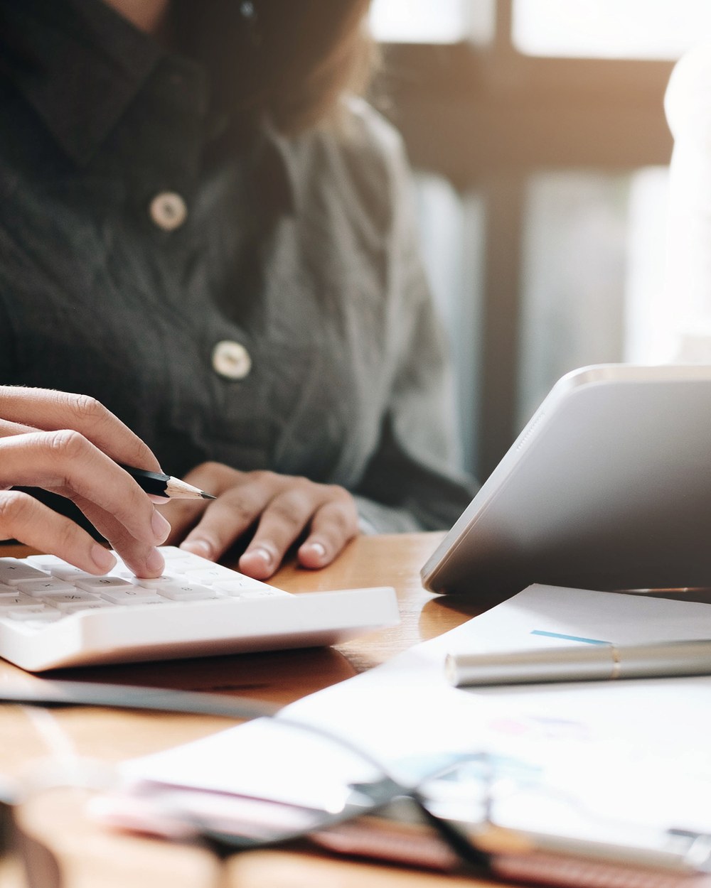 Hands at a desk — calculator, tablet, notebook with charts — warm daylight